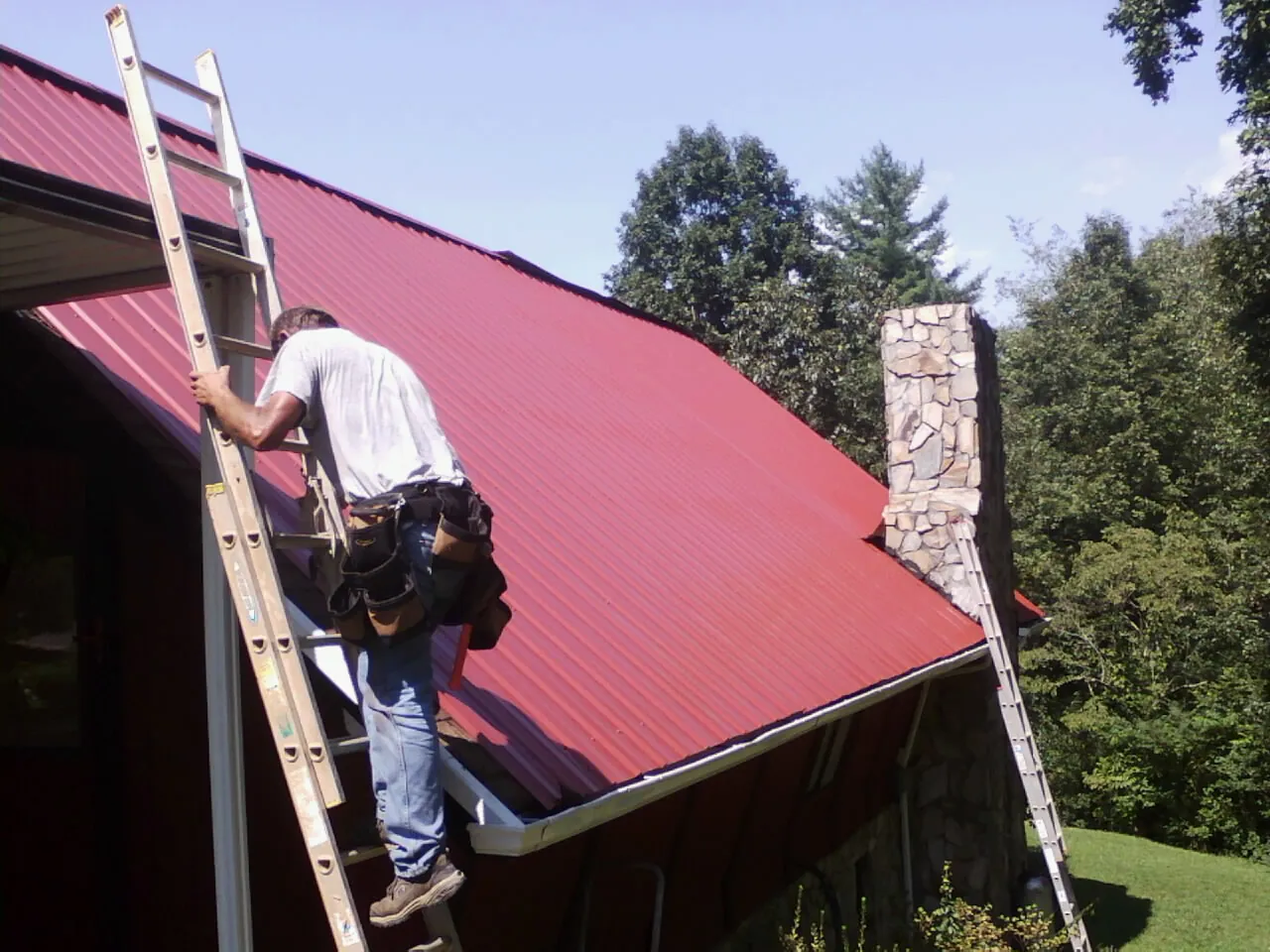 Roofer on a ladder beside a red metal roof and stone chimney — Soffit & Fascia Repair by Roofing Brothers in Roy